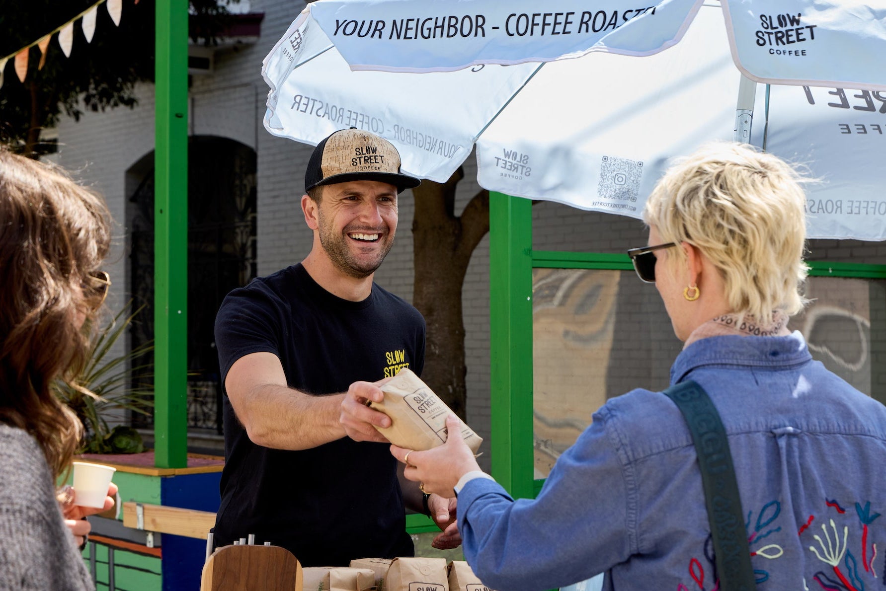 Man handing a coffee bag to a woman under a coffee shop umbrella.